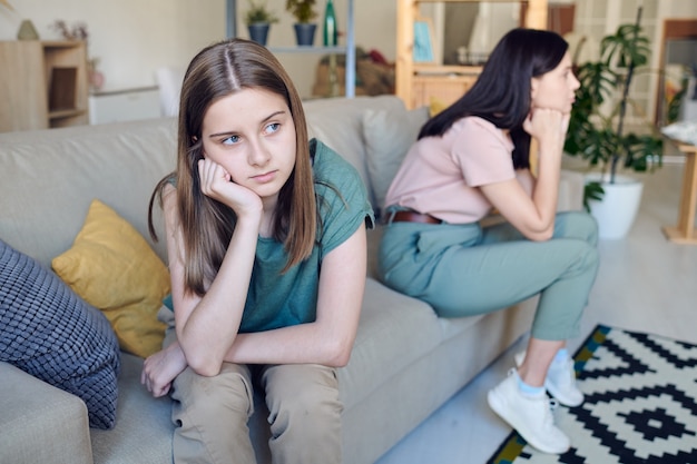 Upset or offended teenage girl with her hand by cheek sitting on couch with her bored or vexed mother during conflict