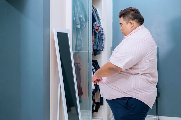 Photo upset overweight asian man wearing tight shirt in front of mirror at home