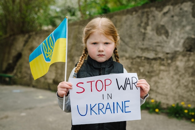 Расстроенная бедная малышка, протестующая против военного конфликта, поднимает баннер с текстом сообщения «Остановить войну в Украине» и сине-желтый флаг «Остановить российскую агрессию» ребенок против войны Нет войне
