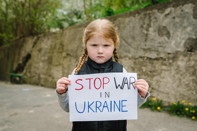 Photo upset poor toddler girl kid protesting war conflict raises banner with inscription message text stop war in ukraine crisis peace stop russian aggression child against war no war stop war