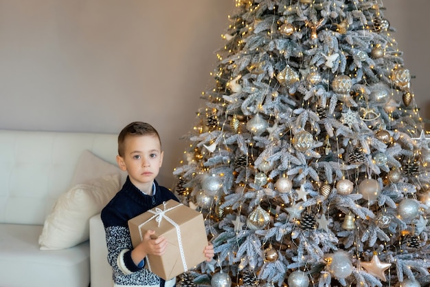 Bambino caucasico in età prescolare turbato con la scatola presente nelle sue mani vicino all'albero di natale. ritratto di ragazzo frustrato la mattina di natale. il bambino ha ricevuto un regalo di natale.