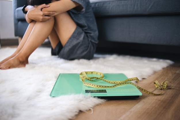 Upset and sad Asian woman sitting on fur rug near weigh scale with measuring tape