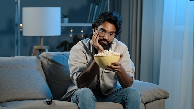 Upset sad bored Arab man tired indian male with beard sits home evening watching boring TV program