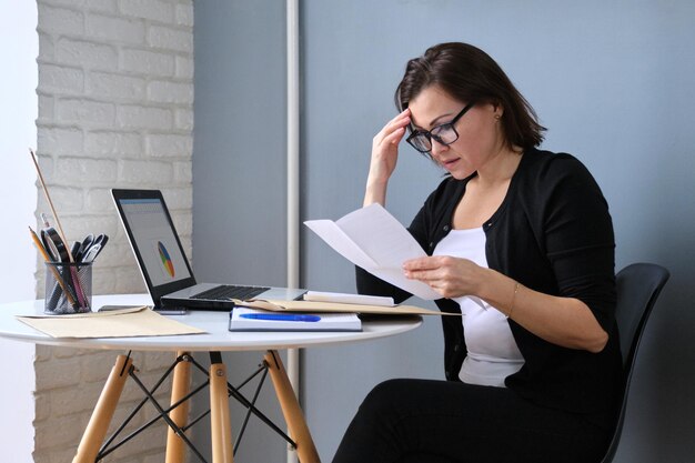 Upset sad middle-aged woman with papers laptop computer sitting at home at the table. Bad read news, financial difficulties, debt pay, payment information