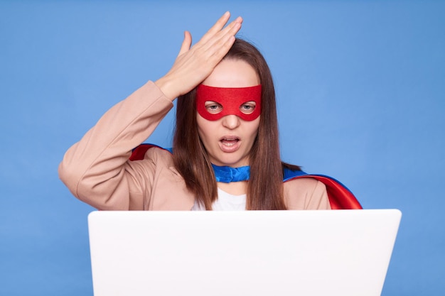 Фото Расстроенная, грустная женщина в костюме супергероя, стоящая в изоляции на синем фоне, работающая на ноутбуке, имеющая проблемы с работой, показывая жест фацепальма