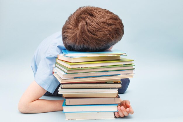 Upset schoolboy sitting with pile of school books boy sleeping on a stack of textbooks