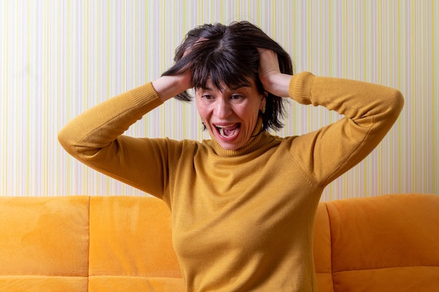 Donna di mezza età urlante sconvolta che soffre di stress tenendo la testa con le mani su un divano arancione crisi di mezza età concetto di salute mentale