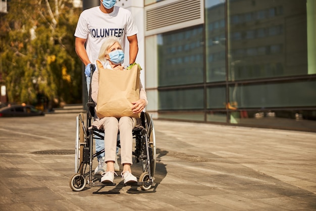 Расстроенная старшая женщина, сидящая в инвалидной коляске и держащая на коленях пакет с продуктами