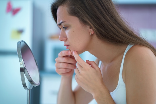 Upset stressed sad acne woman with problem skin squeezes pimple at home in front of a small round mirror