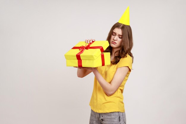 Upset teenager girl opening birthday gift box and looking inside with disappointed expression unwrapping bad present wearing yellow Tshirt and birthday cone Indoor isolated on gray background