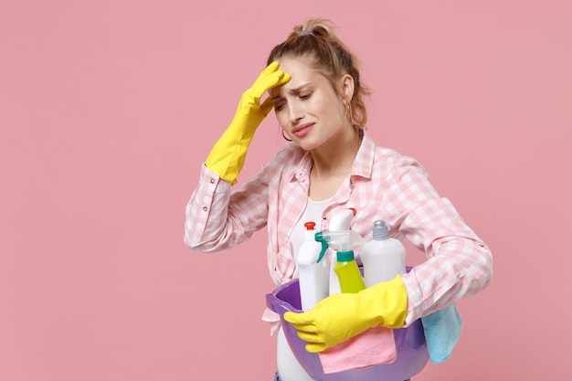 Upset tired young woman housewife in rubber gloves hold basin with detergent bottles washing cleansers while doing housework isolated on pink background. Housekeeping concept. Put hand on head crying.