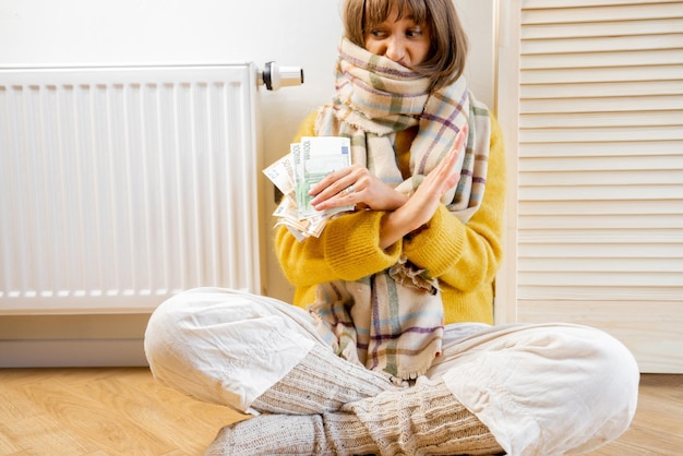 Upset warmly dressed woman with money near heater at home