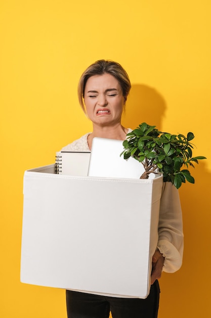 Upset woman holding box with personal items after job resignation against yellow background