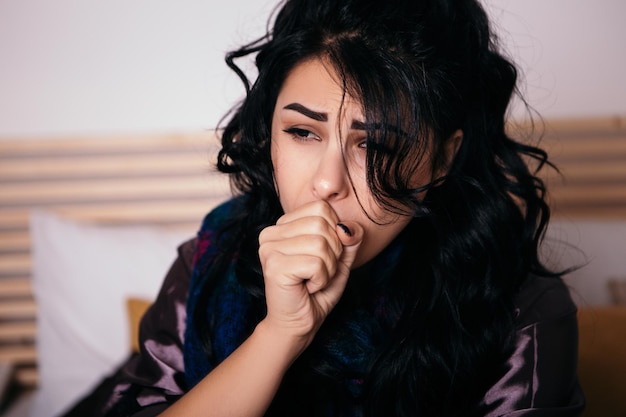 Upset woman lying in bed with scarf and coughing at home