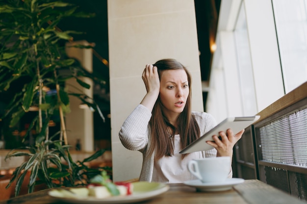 Photo upset woman sitting alone near big window in coffee shop with cup of cappuccino, cake, relaxing during free time. female working, reading bad news on pc tablet computer rest in cafe. lifestyle concept