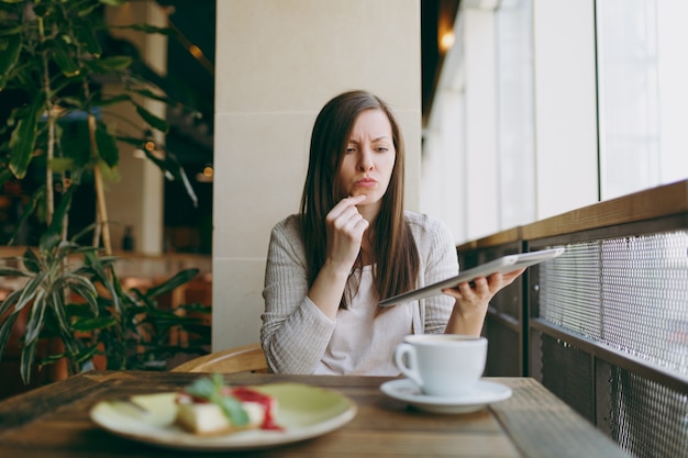 Upset woman sitting alone near big window in coffee shop with cup of cappuccino, cake, relaxing during free time. Female working, reading bad news on pc tablet computer rest in cafe. Lifestyle concept