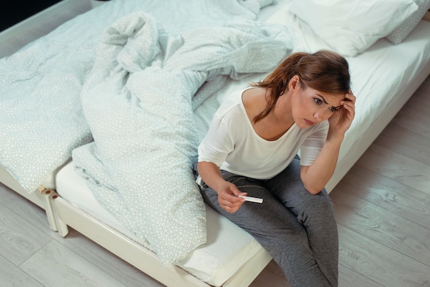 Upset woman sitting sad in white bed with negative pregnancy test