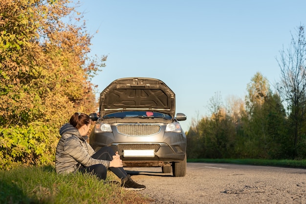 An upset woman stands near her broken car and talking on a cell phone on the road in autumn.