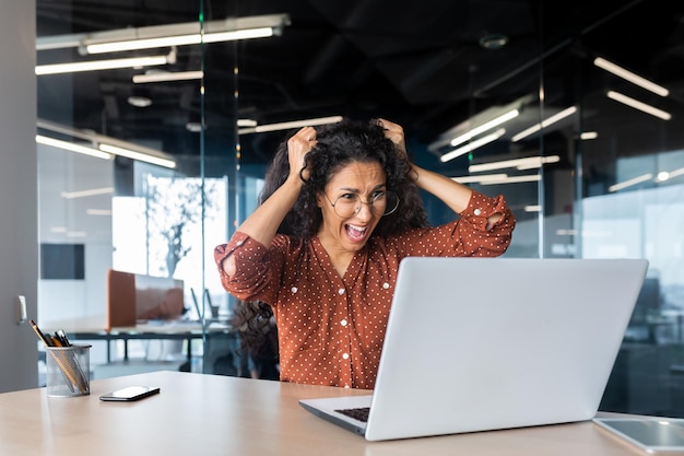 Расстроенная женщина на работе кричит на монитор ноутбука, разъяренная деловая женщина держит волосы на голове в офисе