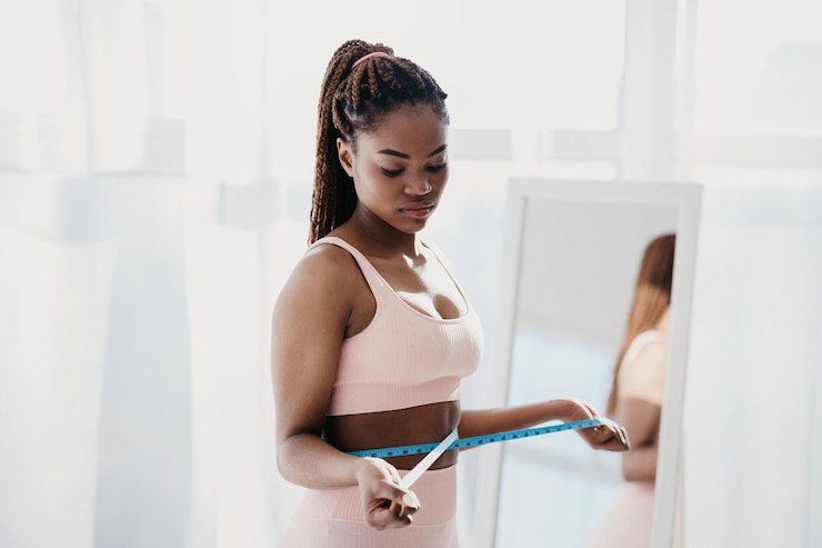Photo upset young african american woman measuring her waist near mirror feeling unhappy with results of