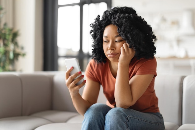 Upset young african american woman sitting on couch and looking at smartphone