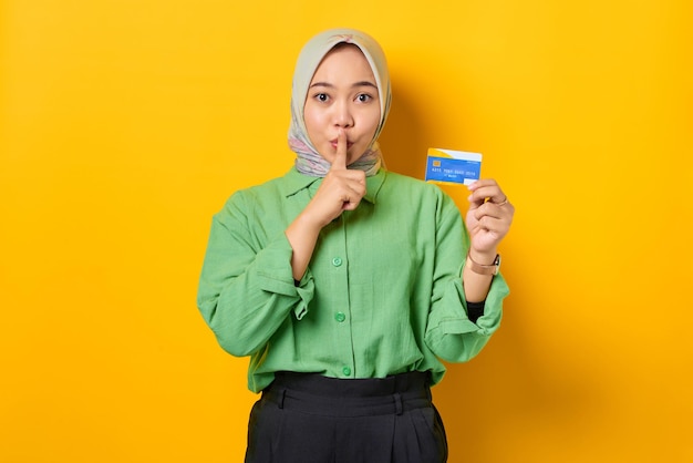 Upset young Asian woman in a green shirt holding credit card and makes silence gesture on yellow background