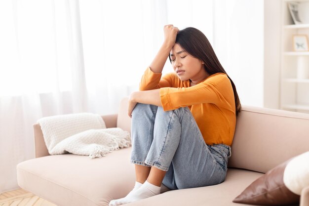 Upset young asian woman sitting on couch touching her head