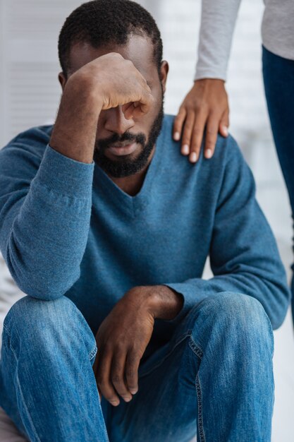 Upset. Young emotional man sitting with his eyes closed and looking depressed while his girlfriend touching his shoulder