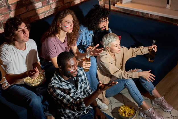 Upset young friends worry about football team in competition, watching sport game on tv, gathered together at home at night, guys and ladies emotionally gesticulating