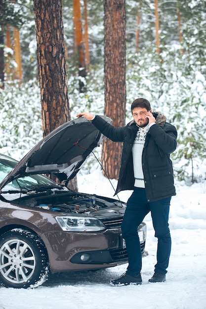 Расстроенный молодой человек звонит в автосервис, стоя на разбитой машине зимой в лесу. машина помощи на дороге