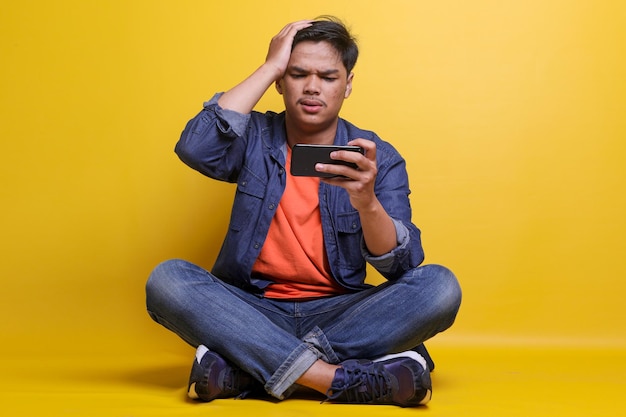 Upset young man looking to mobile phone having confused expression while sitting on yellow color bac