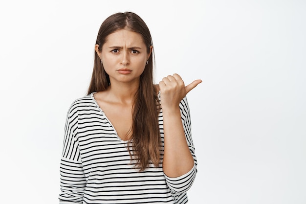 Upset young woman complains, pointing finger right and furrow eyebrows disappointed, sulking of smth upsetting, white background.