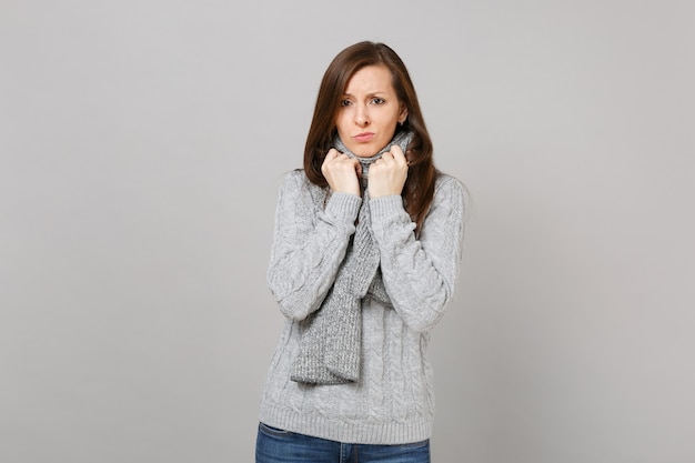 Upset young woman in gray sweater holding hands on scarf isolated on grey wall background, studio portrait. Healthy fashion lifestyle, people sincere emotions, cold season concept. Mock up copy space.