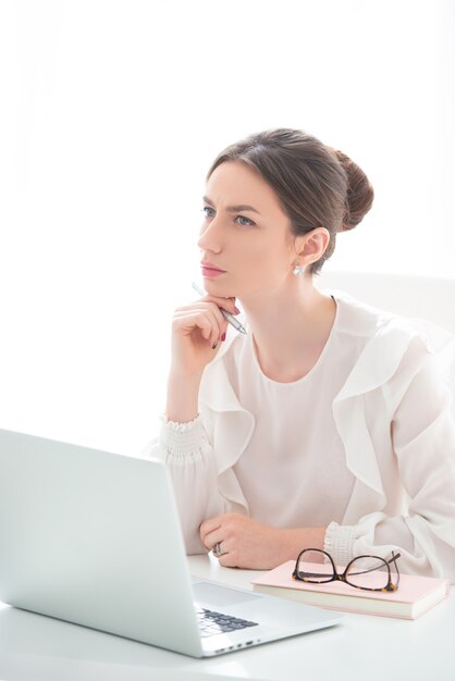 An upset young woman is sitting in an office at a laptop, and is holding her head in her hands. 