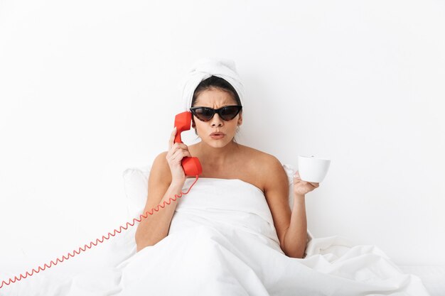 Upset young woman sitting in bed after shower wrapped in blanket, wearing sunglasses, talking on a landline phone, holding a cup