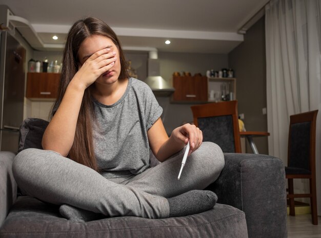 upset young woman sitting on sofa with pregnancy test in hands