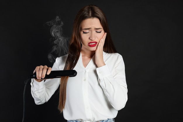 Upset young woman with flattening iron on black background Hair damage