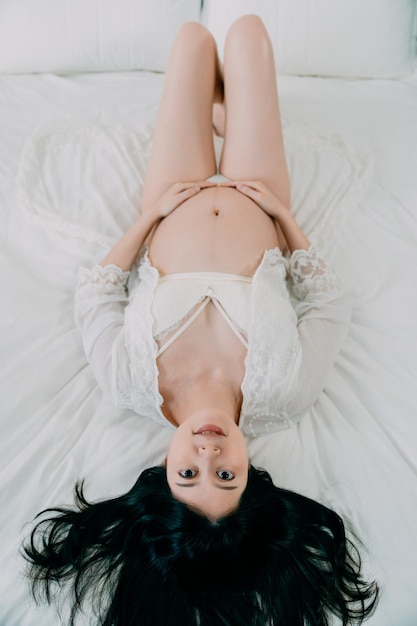 upside down photo of beauty sexy pregnant asian japanese woman in lingerie and white lacy gown lying in cozy bed in bright room. beautiful future mom model smiling face camera with bare belly.