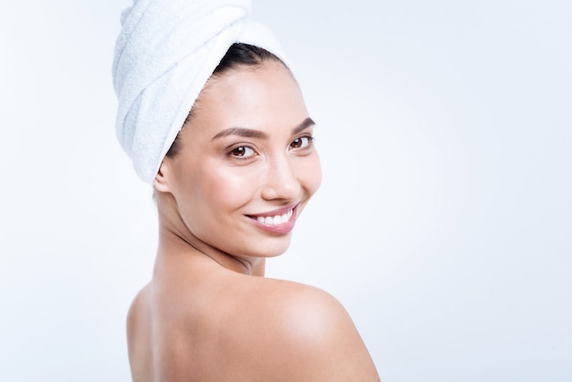 Upsurge of great mood. Beautiful young woman in a towel turban standing half-turned and smiling , having taken a shower