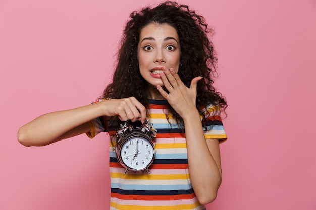 uptight woman 20s with curly hair holding alarm clock isolated on pink