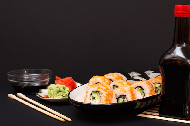 Uramaki California. Sushi rolls with nori, rice, pieces of avocado, cucumber, decorated with flying fish roe on ceramic plate. Bottle and bowl with soy sauce. Plate with red pickled ginger and wasabi.