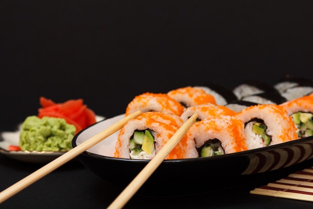 Uramaki California. Sushi rolls with nori, rice, pieces of avocado, cucumber, decorated with flying fish roe on ceramic plate with wooden sticks. Plate with red pickled ginger and wasabi.