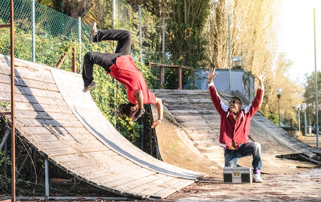 Urban athlete breakdancer performing somersault jump flip at skate park - Afroamerican guy watching friend acrobat dancing with extreme flipping move - Breakdance acrobatic and friendship concept