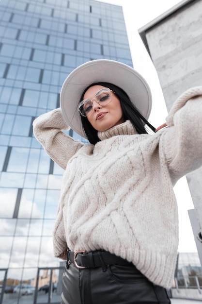 Urban female portrait of beautiful fashion woman model with\
stylish glasses in vintage cozy sweater and white hat travels in\
the city