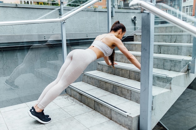 Allenamento urbano della donna di forma fisica che fa piedi sollevati push up sulle scale del parco urbano. atleta femminile motivato che si allena duramente.