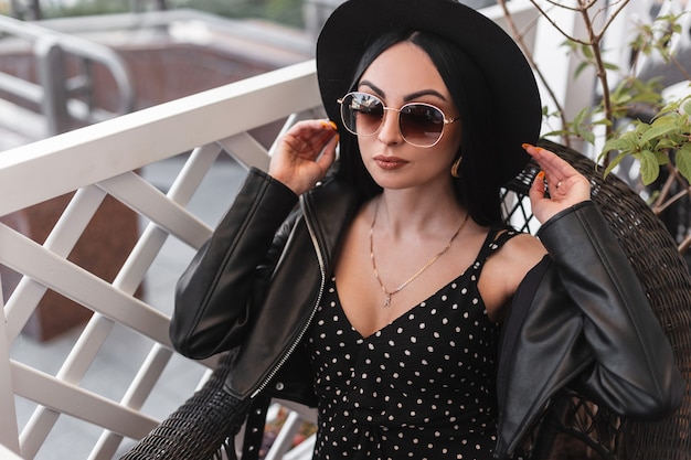 Urban gorgeous woman in fashionable sunglasses in beautiful black dress in leather jacket straightens retro hat. Lovely fashion model girl in black trendy outfit is resting on vintage chair in cafe.