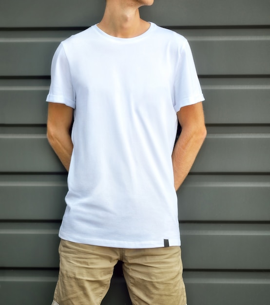 Городской макет одежды. Молодой человек, прислонившись к серой фактурной стене в пустой футболке и коричневых шортах. Шаблон готов для вашего дизайна.