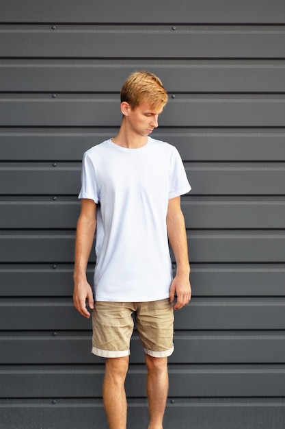 Urban mockup of clothes. Young man  standing  near the gray  metal profiled wall in a white T-shirt and brown shorts.  Template  can use for you design.