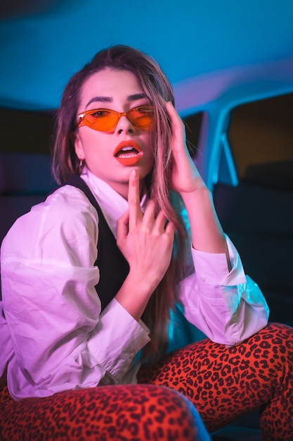 Urban photography with blue and purple neons in a car. Young blonde Caucasian model pose with hearts glasses and a white shirt