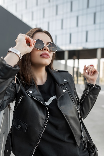 Urban portrait of fashion woman in stylish black clothes with leather jacket and dress puts on fashionable sunglasses in the modern city